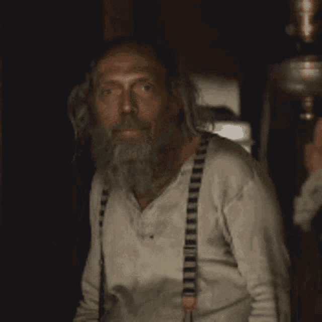 a man with a beard and suspenders is looking at the camera .