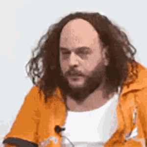 a man with long hair and a beard is wearing an orange jacket and smoking a cigarette .