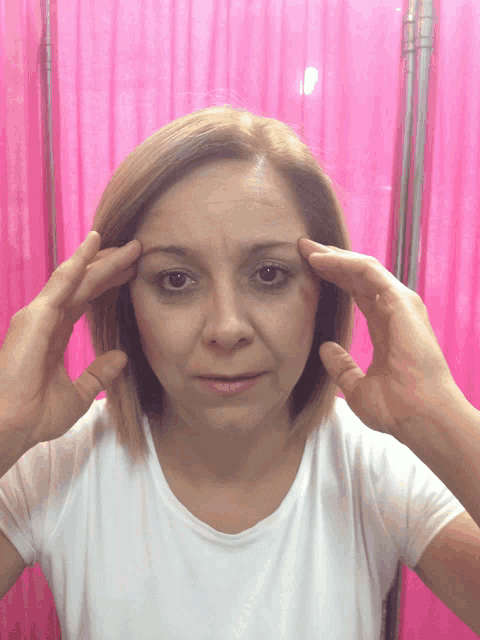 a woman in a white shirt is holding her head in front of a pink curtain
