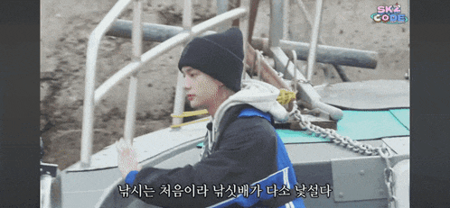 a man wearing a black beanie and a blue jacket is standing on a boat with the words skz code on the bottom right