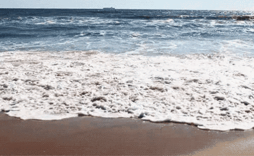 waves crashing on a sandy beach with a large ship in the distance