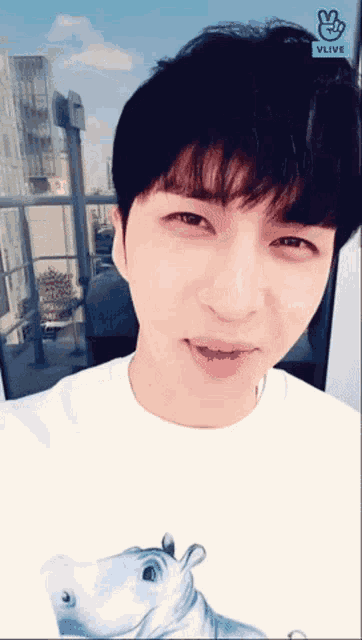 a young man wearing a white shirt with a picture of a hippo on it looks at the camera with a vlive logo visible