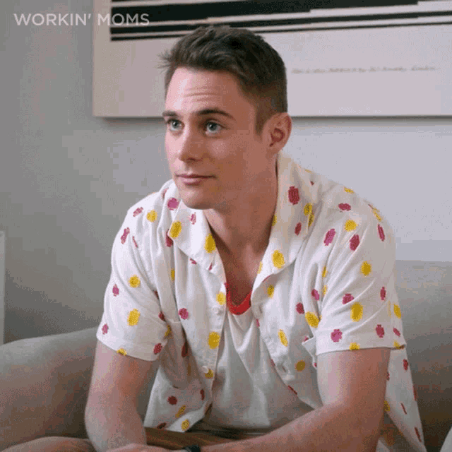 a man in a polka dot shirt sits on a couch in front of a workin ' moms poster