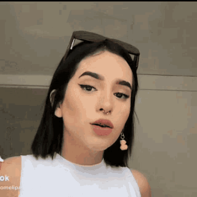 a woman wearing a white tank top and earrings with a teddy bear on them is looking at the camera .