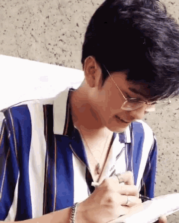 a man wearing glasses and a blue and white striped shirt is writing on a piece of paper .