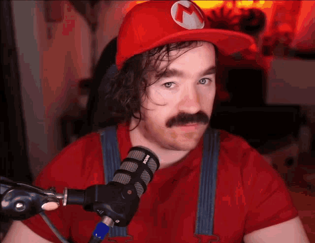 a man with a mustache wearing a red hat and overalls stands in front of a rode microphone