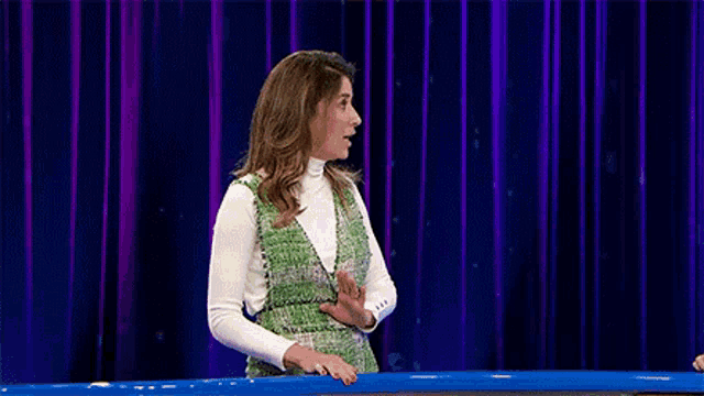 a woman in a green dress and white turtleneck is standing in front of a blue curtain .