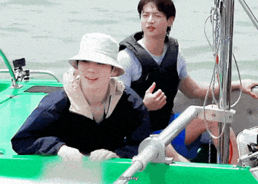 two men are sitting on a green boat and one of them is wearing a bucket hat