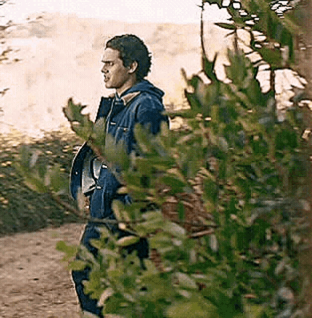 a man in a blue jacket is standing in the woods behind a bush .