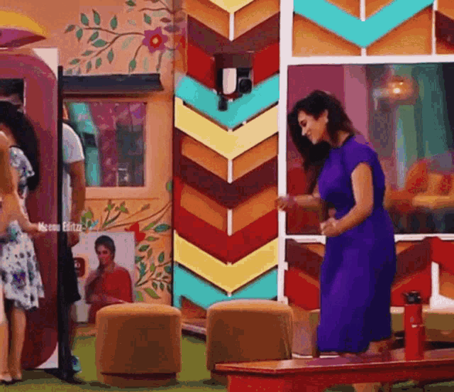 a woman in a purple dress is standing on a wooden bench in front of a colorful wall .