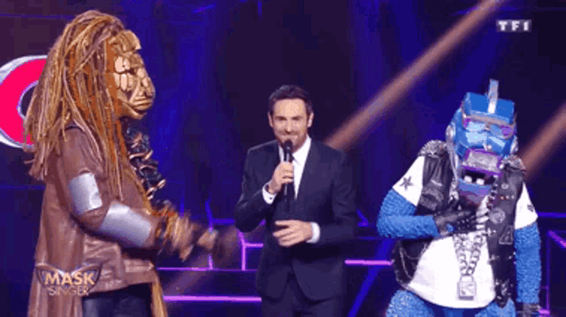 a man in a suit holds a microphone while standing next to two masked people