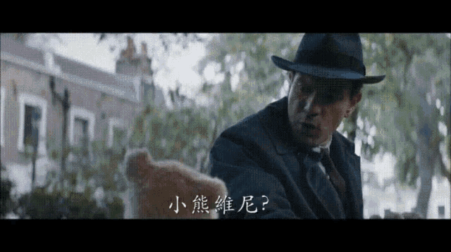 a man in a suit and hat is holding a teddy bear in front of chinese writing