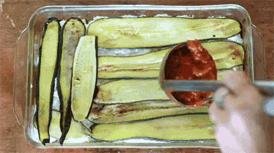 a person is spreading tomato sauce on a casserole dish filled with zucchini slices .