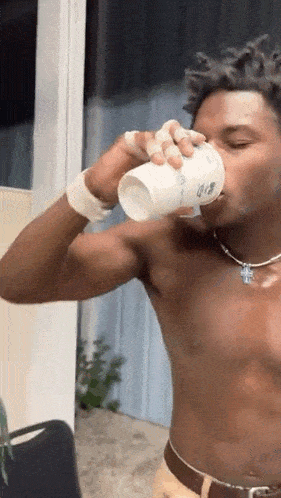 a shirtless man is drinking from a cup while standing in front of a window .