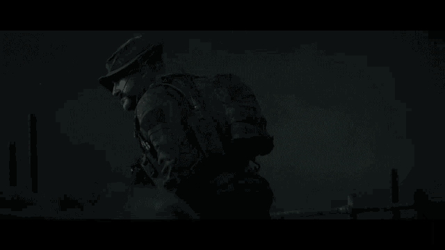 a man in a military uniform is walking in the dark holding a gun and a backpack .