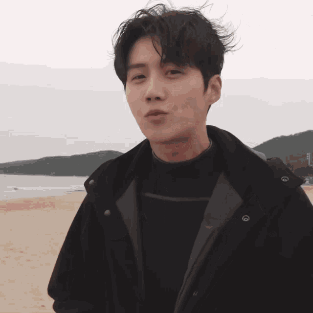 a young man wearing a black jacket stands on a sandy beach
