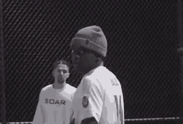 two men are standing next to each other in a cage . one of the men is wearing a soar shirt .