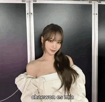 a woman is standing in front of a black wall wearing a white off the shoulder sweater .