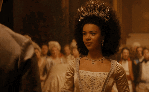 a woman wearing a tiara and a necklace stands in front of a crowd of people