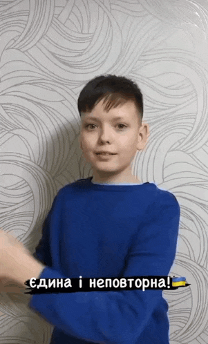 a young boy wearing a blue sweater is standing in front of a wall with a gray pattern
