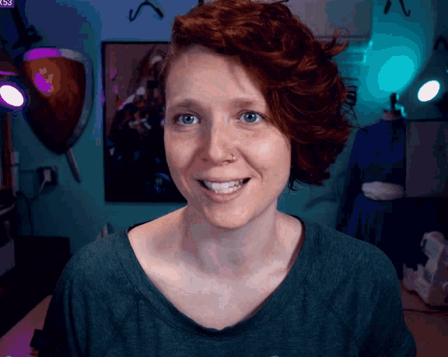 a woman with red hair and a nose ring smiles in front of a blue wall with a shield on it