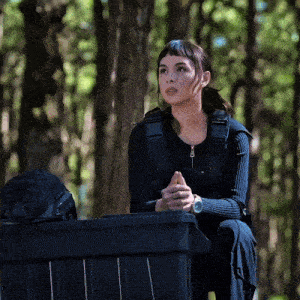 a woman sits on a black bin in the woods with her hands folded
