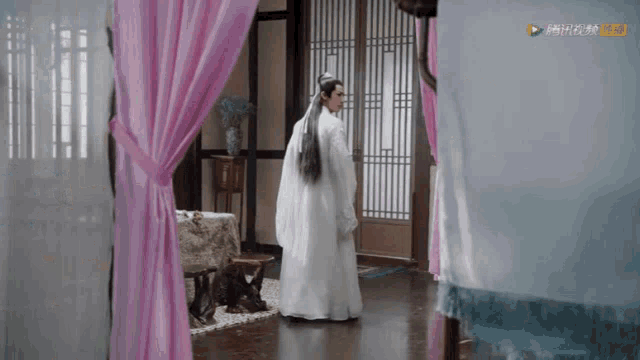 a man in a white dress is standing in a room with pink curtains