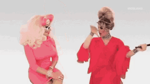 two drag queens , one in a pink dress and one in a red dress , are sitting next to each other .