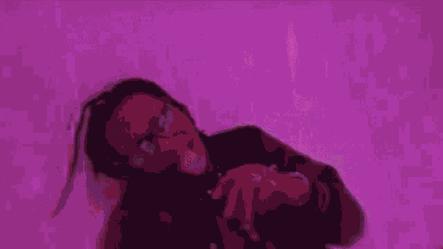 a man with dreadlocks and glasses is laying on a bed in front of a pink background .