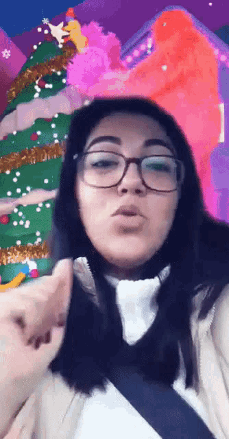 a woman wearing glasses is giving a thumbs up with a christmas tree in the background