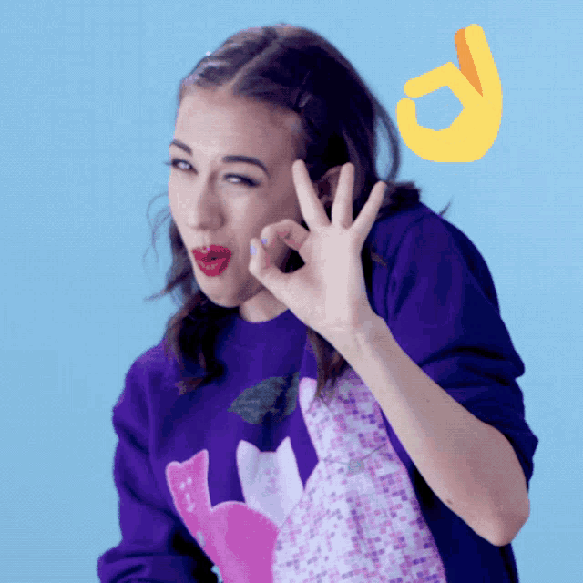 a woman wearing a purple sweater with cats on it gives an ok sign