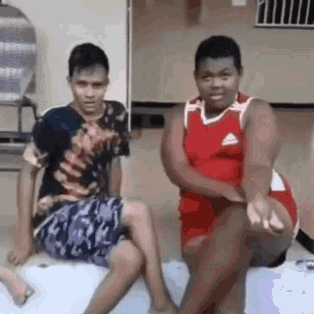 two young men are sitting next to each other on the floor . one of the men is wearing a red adidas shirt .