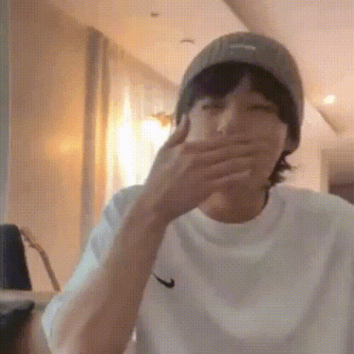 a young man wearing a hat and a white shirt is covering his mouth with his hand .