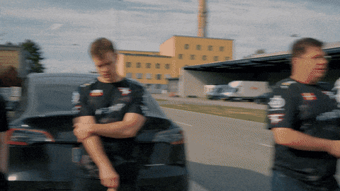 two men standing in front of a car with one wearing a t-shirt that says ' rs ' on it