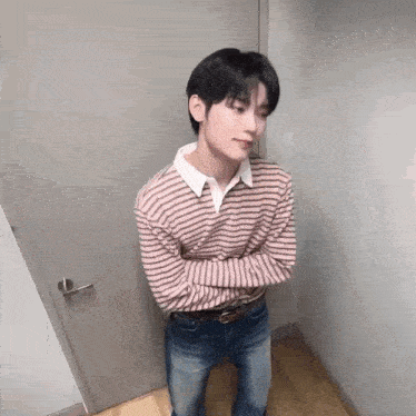 a young man in a striped sweater and jeans is standing in a corner with his arms crossed .