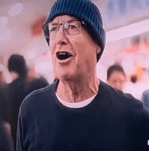 an older man wearing a blue beanie and glasses is standing in a crowded area .