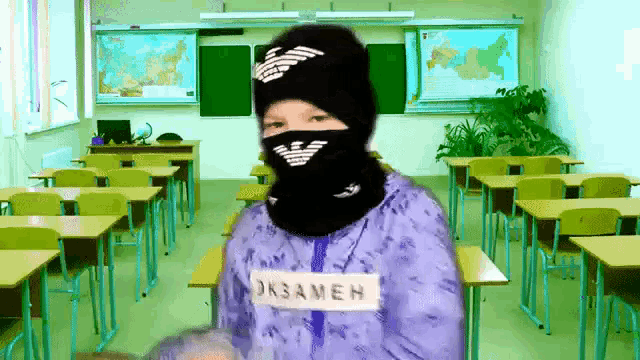 a person wearing a mask and a shirt that says oksameh stands in a classroom