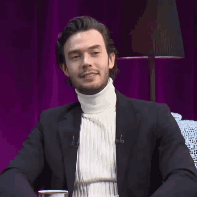 a man wearing a black jacket and a white turtleneck is sitting in front of a lamp and a purple curtain