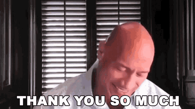 a bald man is smiling and saying thank you so much in front of a window with blinds .
