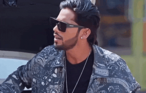 a man with a beard wearing sunglasses and a necklace is standing next to a car .