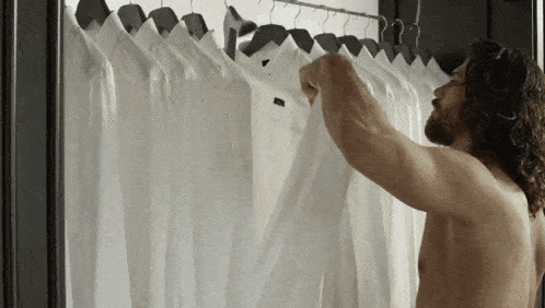 a shirtless man looks at a row of white shirts