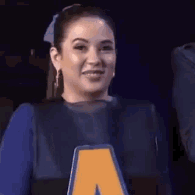 a woman in a blue shirt with a yellow letter a on her chest is smiling .