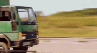 a green truck is driving down a road next to a grassy field .