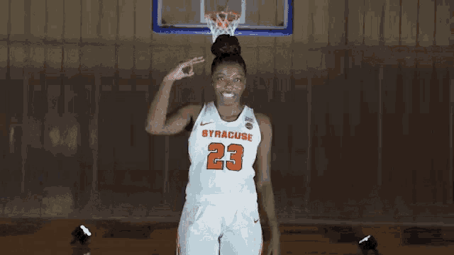 a woman in a syracuse basketball uniform is standing in front of a basketball hoop and giving a peace sign .