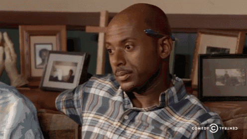 a man in a plaid shirt is sitting on a couch in front of a wall of framed pictures sponsored by central comedy
