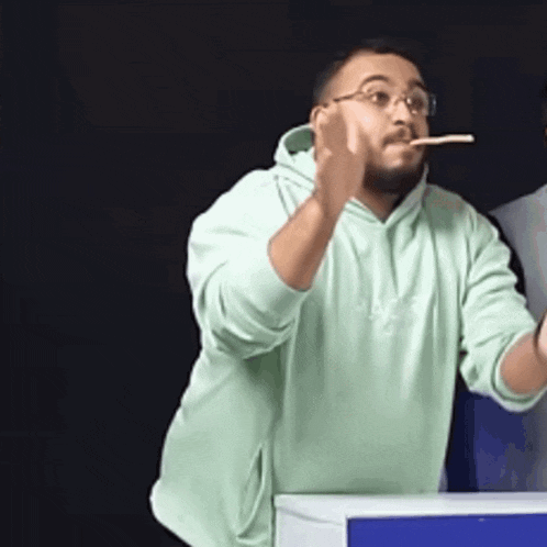 a man wearing glasses and a green hoodie is holding a toothbrush in his mouth
