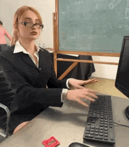 a woman in a suit and glasses is sitting at a desk in front of a computer keyboard .