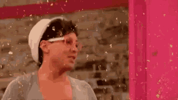 a woman wearing glasses and a white hat is standing in front of a pink door with confetti falling around her .