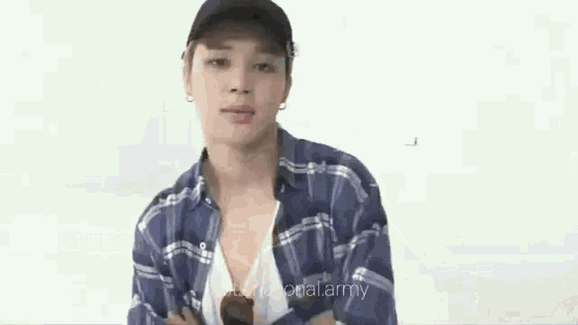 a young man wearing a blue plaid shirt and a black hat is standing in front of a white wall .