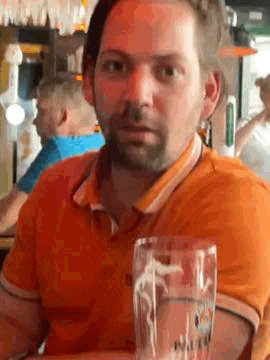 a man in an orange shirt is sitting at a table with a glass of beer in front of him .